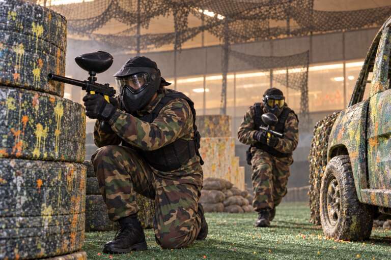 Adult players in camo and full-face masks taking cover behind tyres and a car in the Battle Zone arena