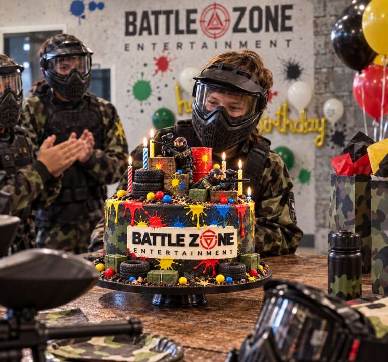 Birthday party at Battle Zone: masked kids around a Battle Zone paintball cake with candles