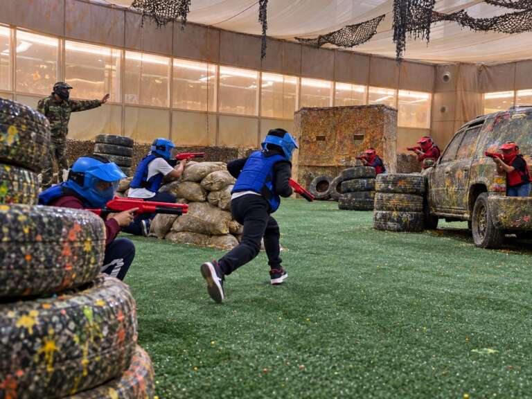 Students in blue and red masks and vests playing a junior paintball game as a Battle Zone marshal guides them